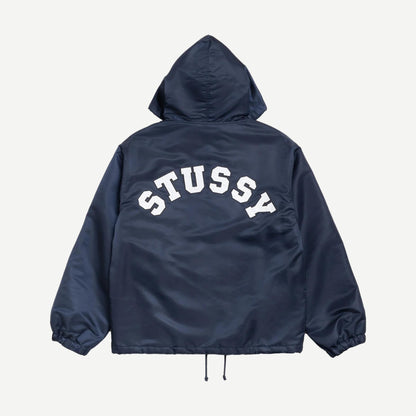 Navy hooded coaches jacket by Stussy, featuring a zip front, adjustable drawstring hood, and ribbed cuffs, displayed on a hanger.