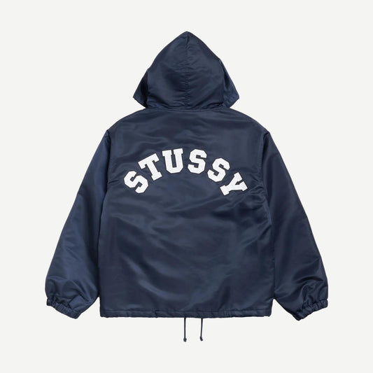 Navy hooded coaches jacket by Stussy, featuring a zip front, adjustable drawstring hood, and ribbed cuffs, displayed on a hanger.