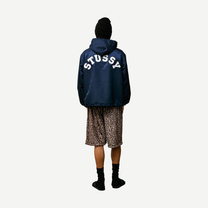 Navy hooded Stussy coaches jacket with front pockets and drawstring hood, displayed on a hanger.